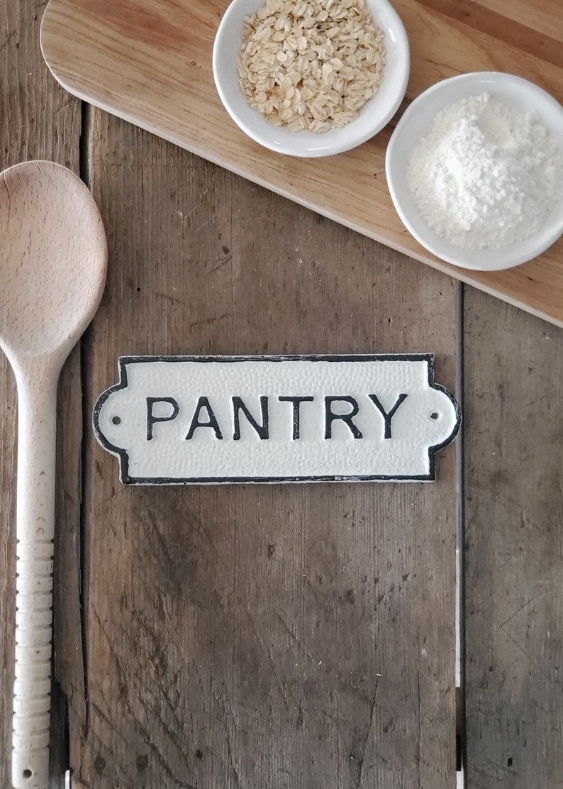 May include: A rustic pantry sign with the word "PANTRY" in black lettering. The sign is white with a black border and is mounted on a wooden surface. A wooden spoon, two small white bowls with oats and flour, and a wooden cutting board are also visible.