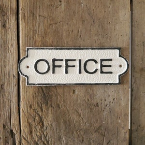 May include: A rectangular, white OFFICE sign with black lettering and a textured surface. The sign has decorative edges and is mounted on a weathered, wooden door. Two small holes are visible on the sign.