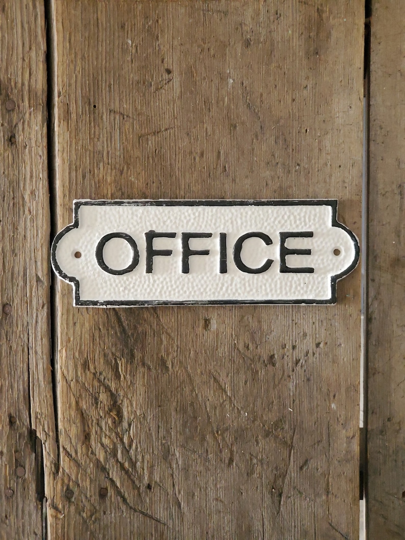 May include: A rectangular, white OFFICE sign with black lettering and a textured surface. The sign has decorative edges and is mounted on a weathered, wooden door. Two small holes are visible on the sign.