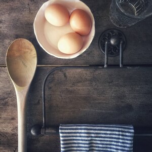 May include: Three brown eggs in a white bowl, a wooden spoon, a striped blue and white tea towel hanging on a black metal rack, and a clear glass bottle on a wooden surface.