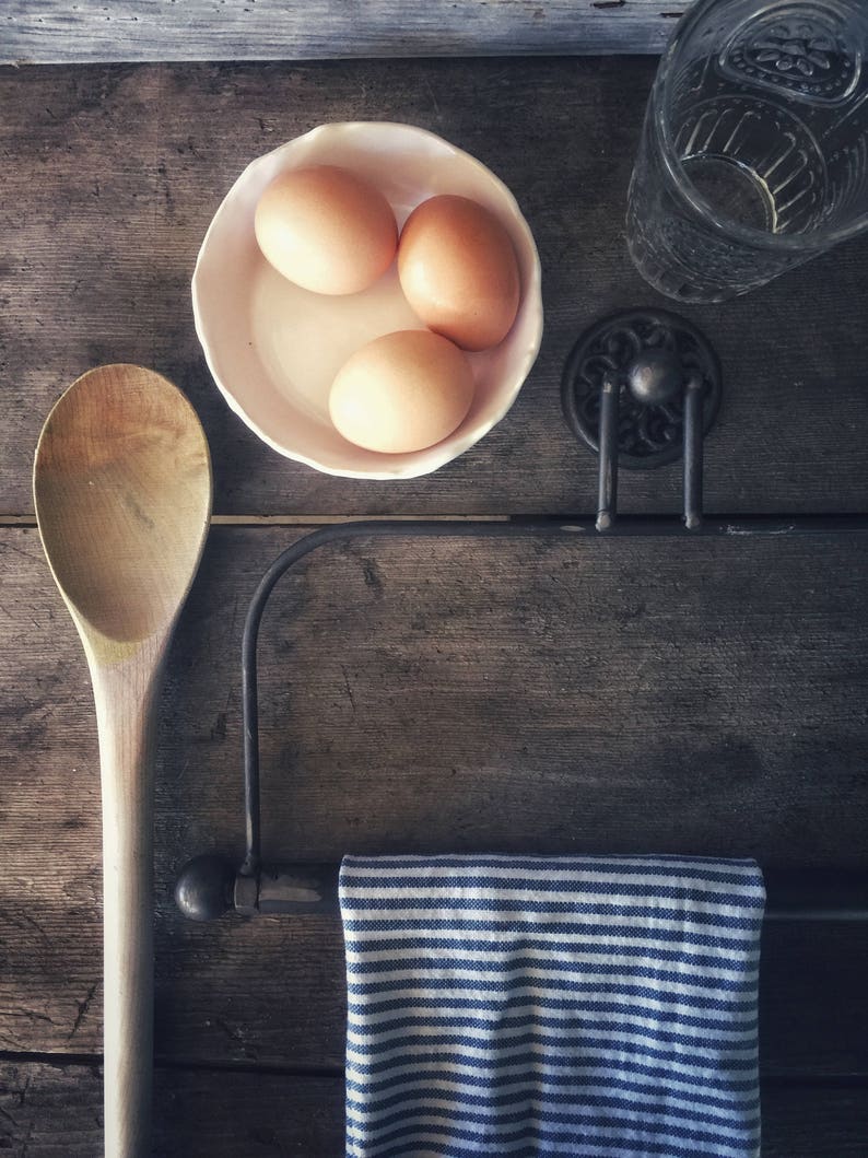 May include: Three brown eggs in a white bowl, a wooden spoon, a striped blue and white tea towel hanging on a black metal rack, and a clear glass bottle on a wooden surface.