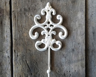 May include: White cast iron hook with a decorative, ornate design. The hook is mounted on a weathered wooden surface.