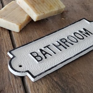 May include: Two bars of handmade soap and a white metal "BATHROOM" sign with black lettering. The soap is light brown with visible plant matter. The sign has a textured surface and a decorative shape.