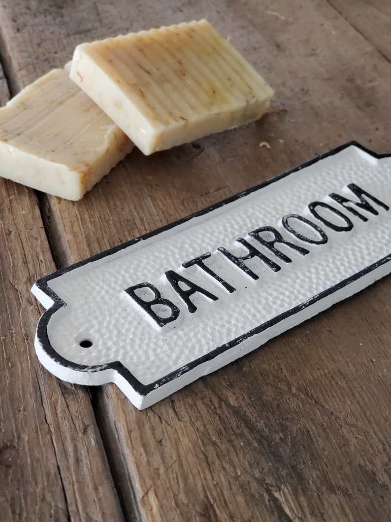 May include: Two bars of handmade soap and a white metal "BATHROOM" sign with black lettering. The soap is light brown with visible plant matter. The sign has a textured surface and a decorative shape.