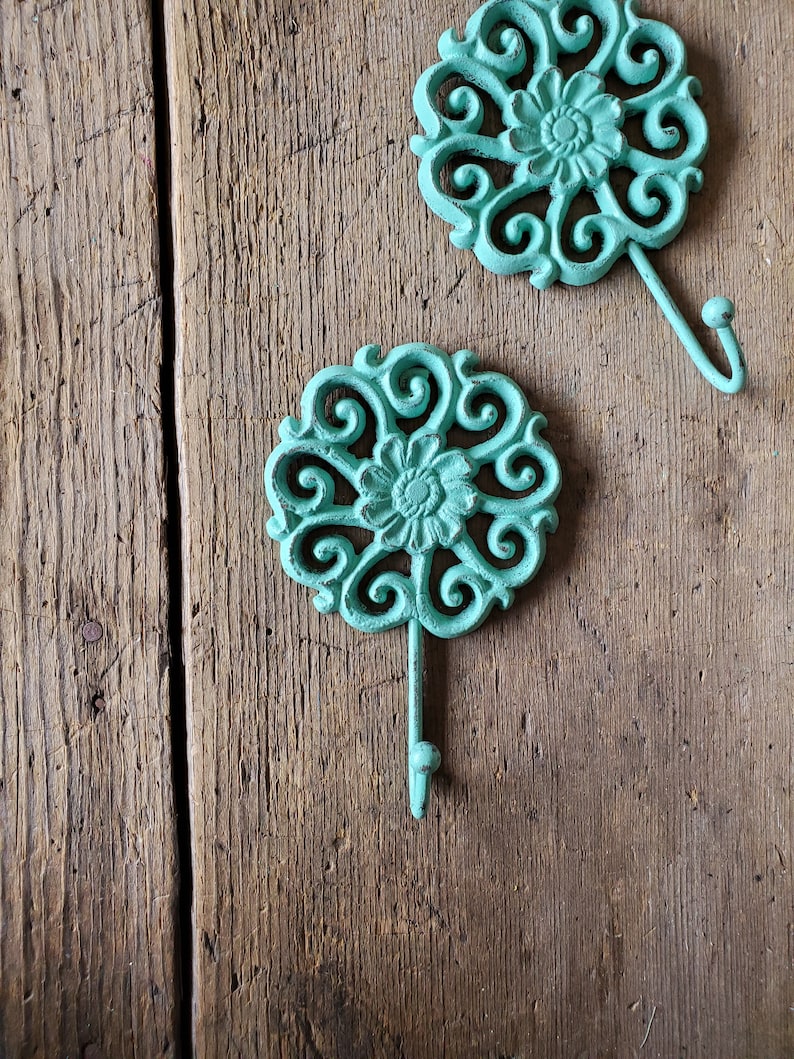 Puede incluir: Dos ganchos de metal azul turquesa con un dise&ntilde;o floral decorativo. Los ganchos est&aacute;n unidos a una superficie de madera.