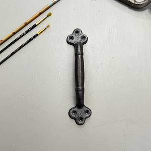 Pode incluir: Puxador de porta de ferro fundido preto com um design decorativo em forma de trevo na parte superior e inferior. O puxador é liso e tem um acabamento ligeiramente texturizado.