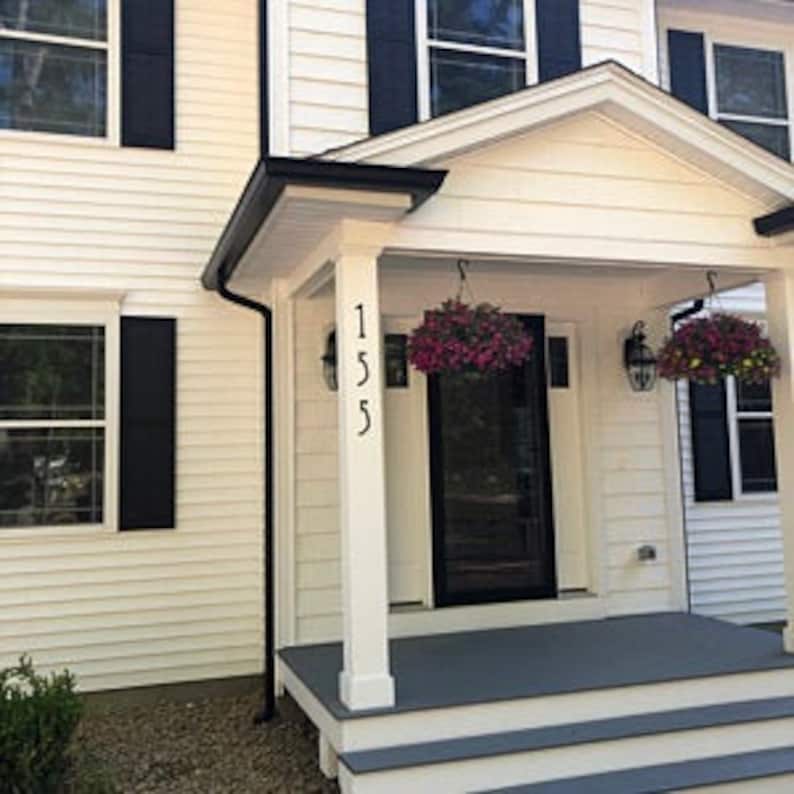 May include: A white house with a black door and black shutters. The house number 155 is displayed on a white column.