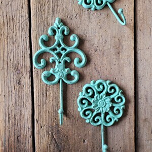 Puede incluir: Tres ganchos decorativos con un acabado azul verdoso. Los ganchos est&aacute;n hechos de hierro fundido y tienen un dise&ntilde;o floral. Est&aacute;n montados en una superficie de madera.
