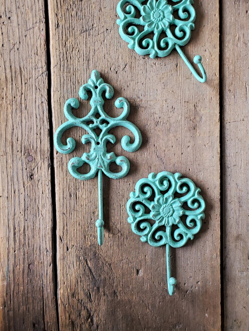 Puede incluir: Tres ganchos decorativos con un acabado azul verdoso. Los ganchos est&aacute;n hechos de hierro fundido y tienen un dise&ntilde;o floral. Est&aacute;n montados en una superficie de madera.
