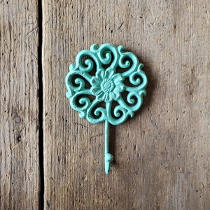 Puede incluir: Un gancho de hierro fundido de color turquesa con un dise&ntilde;o floral decorativo. El gancho est&aacute; montado en una superficie de madera.