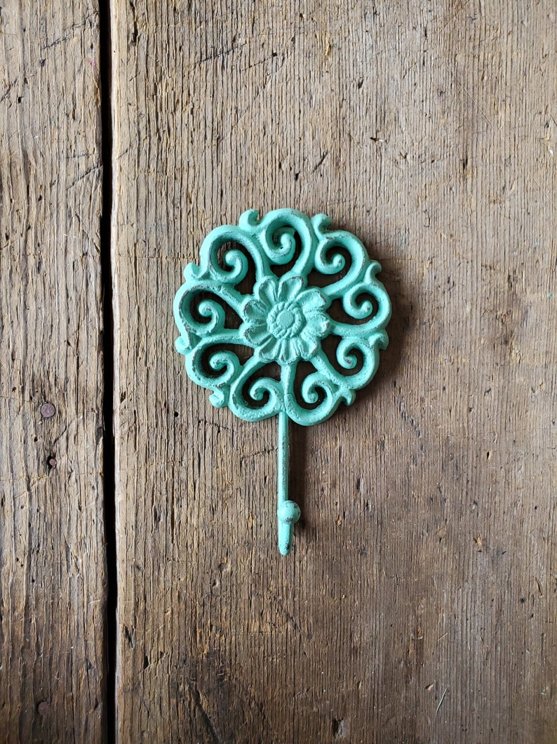 Puede incluir: Un gancho de hierro fundido de color turquesa con un dise&ntilde;o floral decorativo. El gancho est&aacute; montado en una superficie de madera.