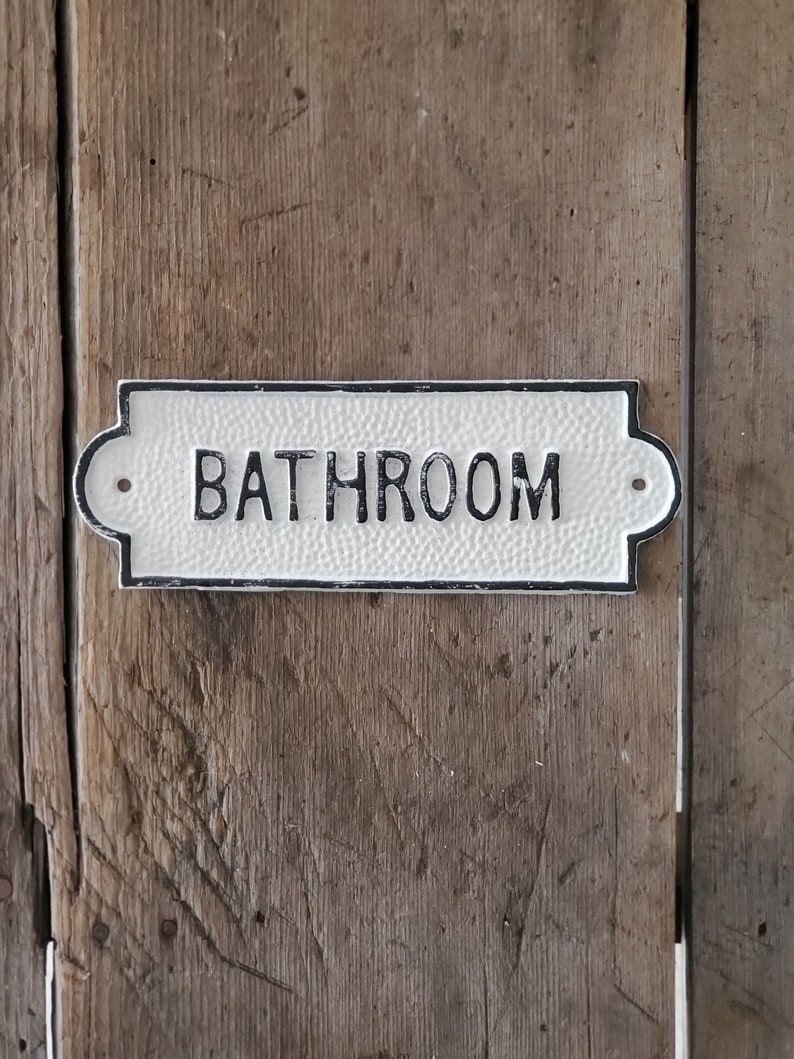 May include: A rectangular, off-white bathroom sign with black lettering and a decorative border. The sign is mounted on a weathered, wooden surface, showcasing a rustic aesthetic. The word "BATHROOM" is prominently displayed.