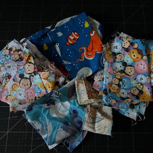 May include: A collection of colorful fabric scraps featuring cartoon characters like Finding Nemo's Dory and characters from the animated film Totoro. The fabrics are cut into various shapes and sizes, ready for a quilting project.