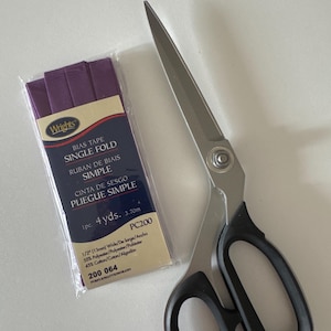 Op de afbeelding: Een pakket paars biaisband en een zilveren en zwarte schaar. Het biaisband is gemarkeerd met "Wright's BIAS TAPE SINGLE FOLD" en is 13 mm breed. De schaar heeft grote zwarte handvatten.