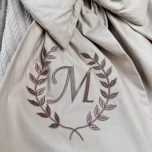 May include: A beige fabric with a large bow and an embroidered wreath with the letter "M" in the center.