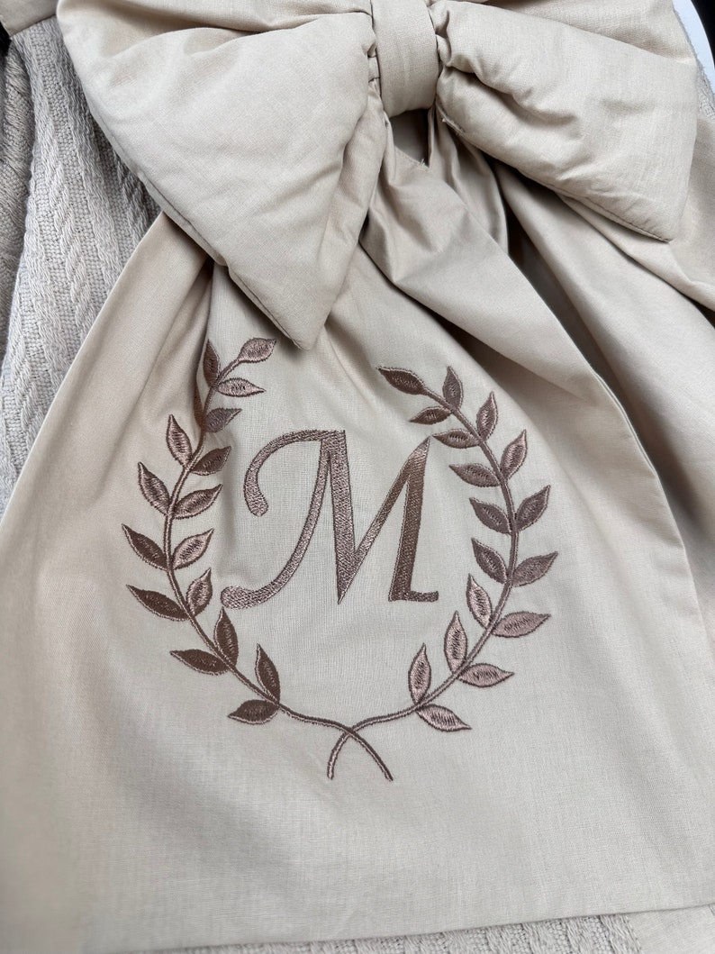 May include: A beige fabric with a large bow and an embroidered wreath with the letter "M" in the center.