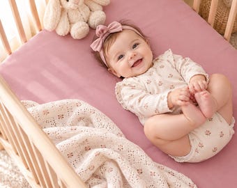 Puede incluir: Un bebé yace sobre una sábana de cuna color rosa empolvado dentro de una cuna de madera. El bebé viste un body color crema con estampado floral y una diadema con lazo a juego. Un conejo de peluche crema y una manta de encaje blanco están cerca.