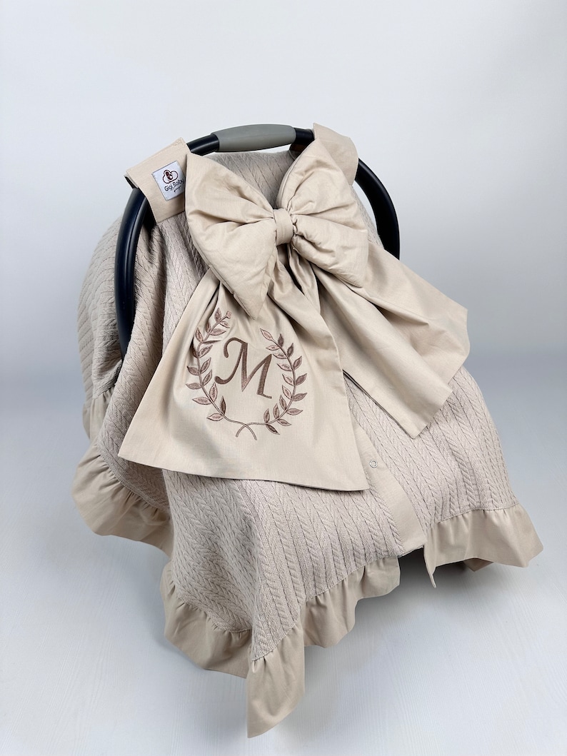 May include: Beige knitted car seat cover with a large beige bow and a monogrammed "M" in a wreath of leaves. The cover has a ruffled edge.