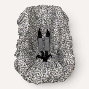 May include: A gray baby car seat cover with a white daisy floral pattern. The cover has black straps and gray padded shoulder straps. The cover is designed to fit over a car seat and protect the baby from the elements.