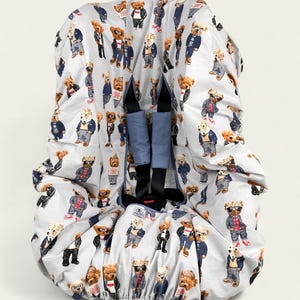 Op de afbeelding: Een lichtgrijze baby autostoelbekleding met een herhalend patroon van teddyberen in verschillende outfits. De beren dragen jassen, pakken en zonnebrillen. De hoes heeft blauwe veiligheidsriemen.