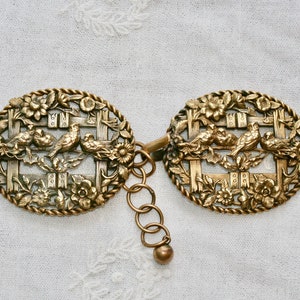 May include: A pair of antique gold-tone metal belt buckles featuring a design of birds in a cage. The buckles are connected by a chain with a small ball at the end.