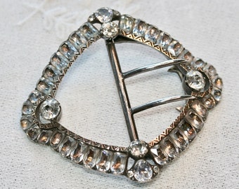 May include: A silver buckle with a decorative design of clear rhinestones. The buckle has a rectangular shape with rounded corners and a central bar with two prongs.