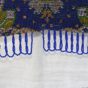 May include: Close-up of a beaded purse with a dark blue background and intricate floral patterns in gold, green, and purple. The purse has a fringe of blue beads along the bottom edge. The background is white.