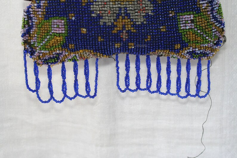 May include: Close-up of a beaded purse with a dark blue background and intricate floral patterns in gold, green, and purple. The purse has a fringe of blue beads along the bottom edge. The background is white.