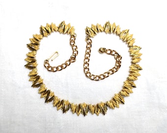 May include: A gold-tone necklace with a chain and a pendant made of small, pointed leaves. The necklace is on a white background.