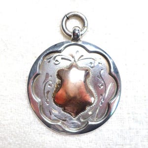 Pode incluir: Pingente prateado com um design decorativo. O pingente apresenta uma forma de escudo central na cor cobre, rodeado por um padrão ornamental em forma de flor. Um pequeno anel é preso no topo para pendurar.