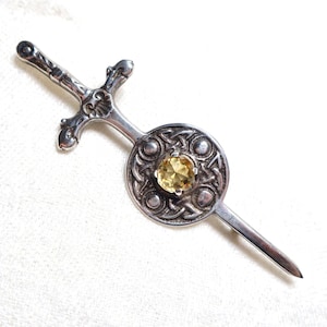 Puede incluir: Broche plateado con forma de espada y escudo. La espada tiene una empuñadura detallada, y el escudo presenta una gema amarilla central. El diseño incorpora nudos celtas, creando una estética vintage.