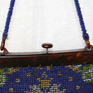 May include: Vintage beaded handbag with a tortoiseshell frame and a beaded blue strap. The bag features a detailed beaded design in blue, gold, green, and white. The clasp is a small, round, dark brown button.