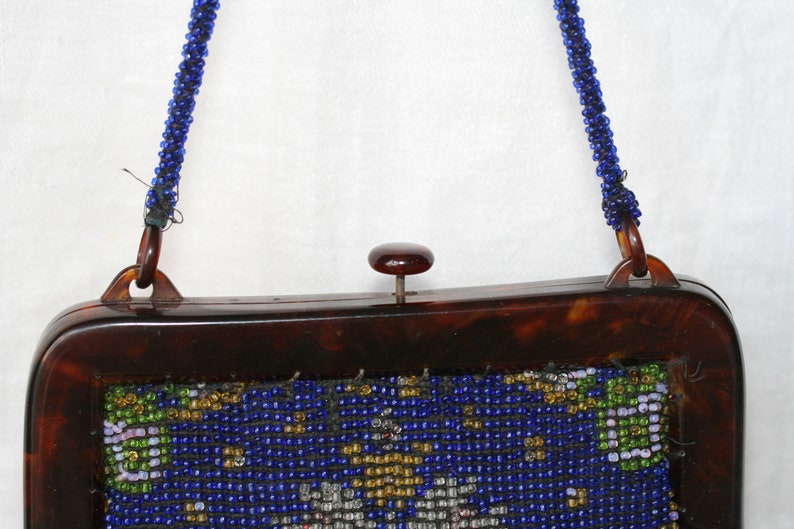 May include: Vintage beaded handbag with a tortoiseshell frame and a beaded blue strap. The bag features a detailed beaded design in blue, gold, green, and white. The clasp is a small, round, dark brown button.