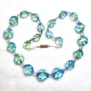 May include: A vintage necklace with faceted, iridescent beads in blue and green hues. The beads are strung with clear beads and a gold-tone clasp. The necklace is displayed on a white surface. The necklace is approximately 40 cm in length.