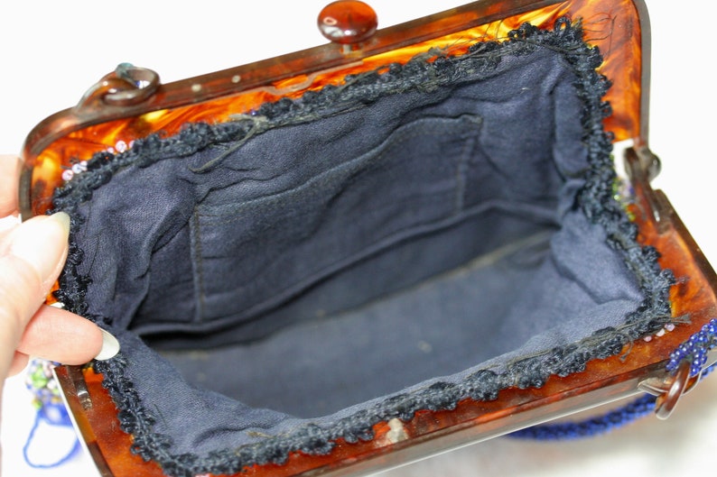 May include: Open view of a vintage handbag with a tortoiseshell frame and a dark blue interior. The frame has a clasp and a beaded handle. The interior is lined with dark blue fabric and has a small pocket.