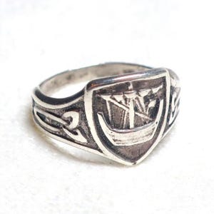 Könnte beinhalten: Silberfarbener Ring mit einem Schilddesign mit Schiffs-Motiv. Der Ring hat eine detaillierte, strukturierte Oberfläche und ein Band mit keltischen Knotenmustern. Der Ring ist wahrscheinlich aus Sterlingsilber und für den täglichen Gebrauch geeignet.