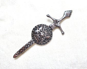 May include: A silver brooch depicting a sword and shield. The shield is circular with an embossed Celtic design. The sword has a detailed handle and a braided blade. The brooch is set against a white background.