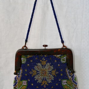 May include: A vintage beaded handbag with a dark blue background and a floral design in gold, white, and green. The purse has a tortoiseshell frame and a beaded handle and fringe.