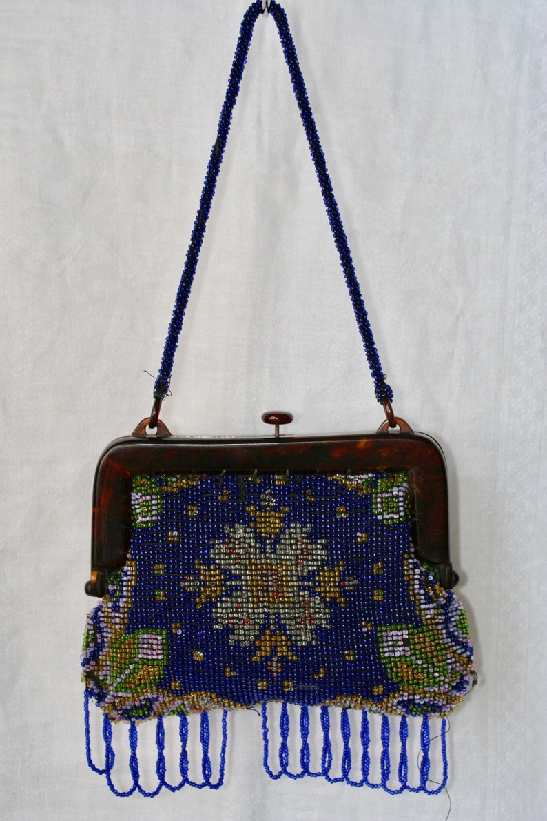 May include: A vintage beaded handbag with a dark blue background and a floral design in gold, white, and green. The purse has a tortoiseshell frame and a beaded handle and fringe.