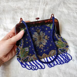 May include: A vintage beaded handbag with a tortoiseshell frame. The bag features a dark blue beaded design with floral accents in white, yellow, and green. The bag has a beaded handle and fringe along the bottom.
