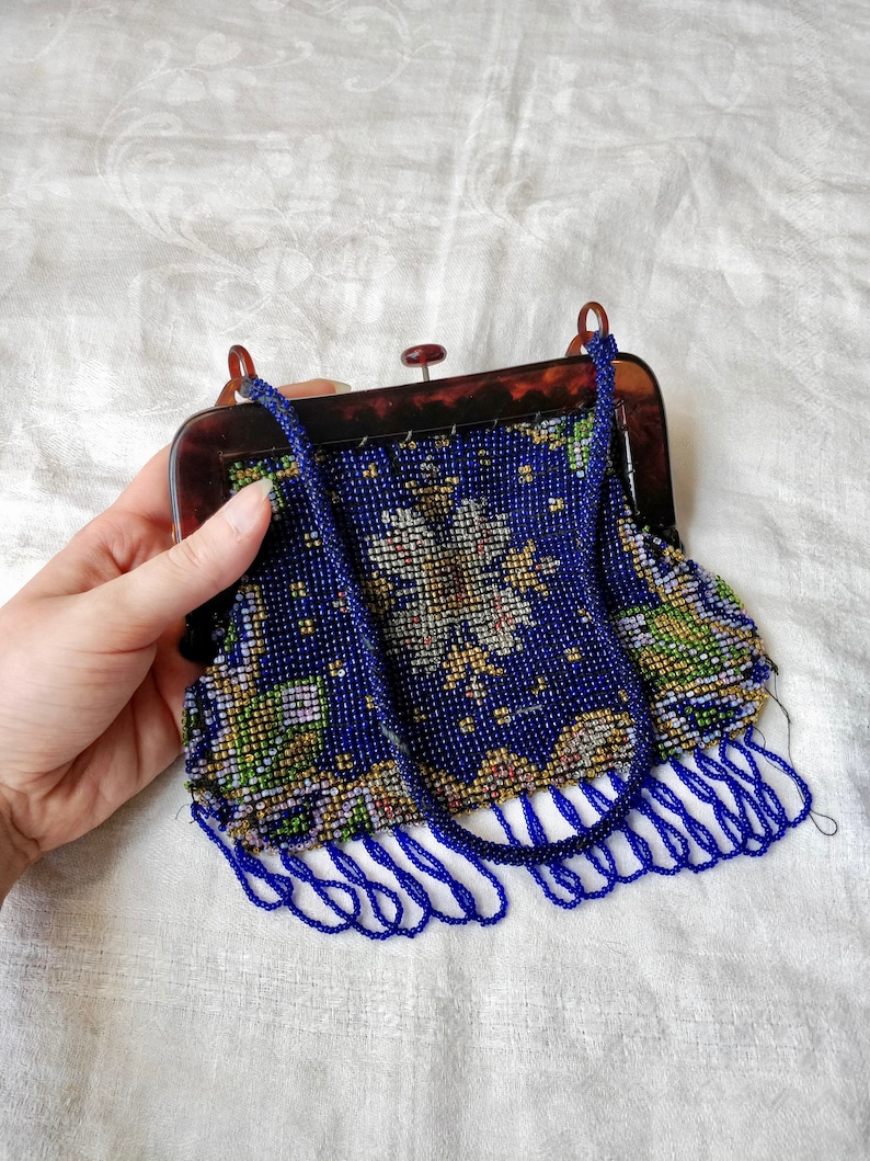 May include: A vintage beaded handbag with a tortoiseshell frame. The bag features a dark blue beaded design with floral accents in white, yellow, and green. The bag has a beaded handle and fringe along the bottom.
