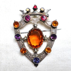 Puede incluir: Broche plateado con diseño de escudo, adornado con una piedra central ovalada de color ámbar y gemas de colores. El broche presenta piedras rojas, moradas, naranjas y azules, y un detalle en forma de corona en la parte superior.