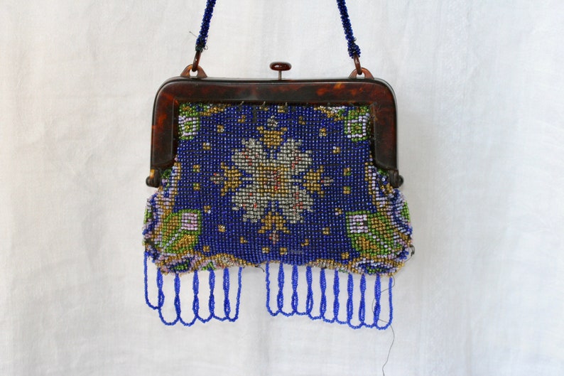 May include: A vintage beaded purse with a dark blue background and a floral design in white, yellow, and green. The purse has a tortoiseshell frame and a beaded handle. Blue beaded fringe hangs from the bottom.