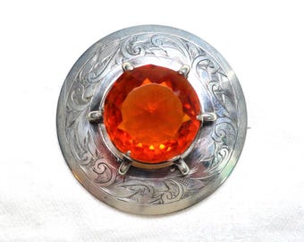 May include: A silver brooch with an orange gemstone center. The gemstone is round and faceted, set in a silver frame with six prongs. The brooch has an engraved floral design around the gemstone.