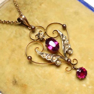 Puede incluir: Un collar de oro con un colgante en forma de corazón que presenta una piedra preciosa rosa y detalles de perlas.