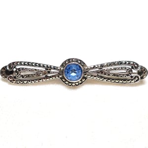 May include: A silver-toned bar pin with a central, round, light blue gemstone. The pin features an ornate design with small, dark accents, likely marcasite stones, and a curved, symmetrical shape.