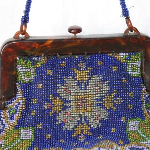 May include: Vintage beaded handbag with a tortoiseshell frame. The bag features a vibrant blue background with a detailed floral pattern in gold, silver, green, and white beads. A beaded strap and a clasp complete the design.