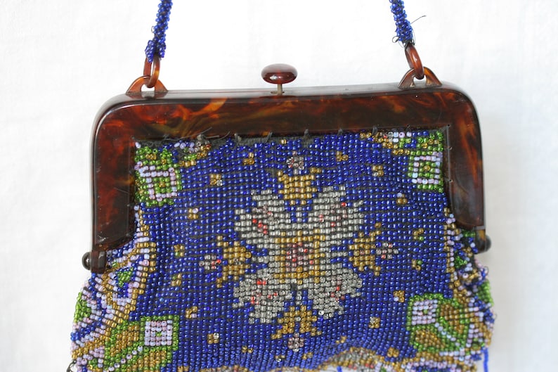 May include: Vintage beaded handbag with a tortoiseshell frame. The bag features a vibrant blue background with a detailed floral pattern in gold, silver, green, and white beads. A beaded strap and a clasp complete the design.