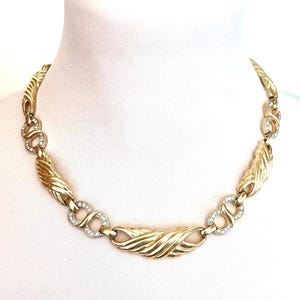May include: Gold-coloured necklace with a textured, wave-like design. The necklace features clear, sparkling accents and a series of interlocking links. The necklace is displayed on a white mannequin.