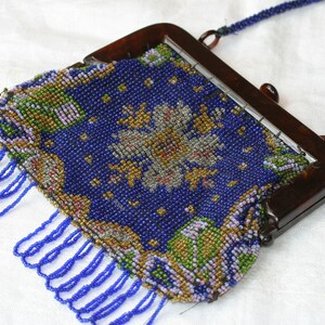 May include: Vintage beaded handbag with a dark brown frame and a beaded strap. The bag features a blue background with a floral design in gold, green, and white beads. Blue fringe hangs from the bottom.